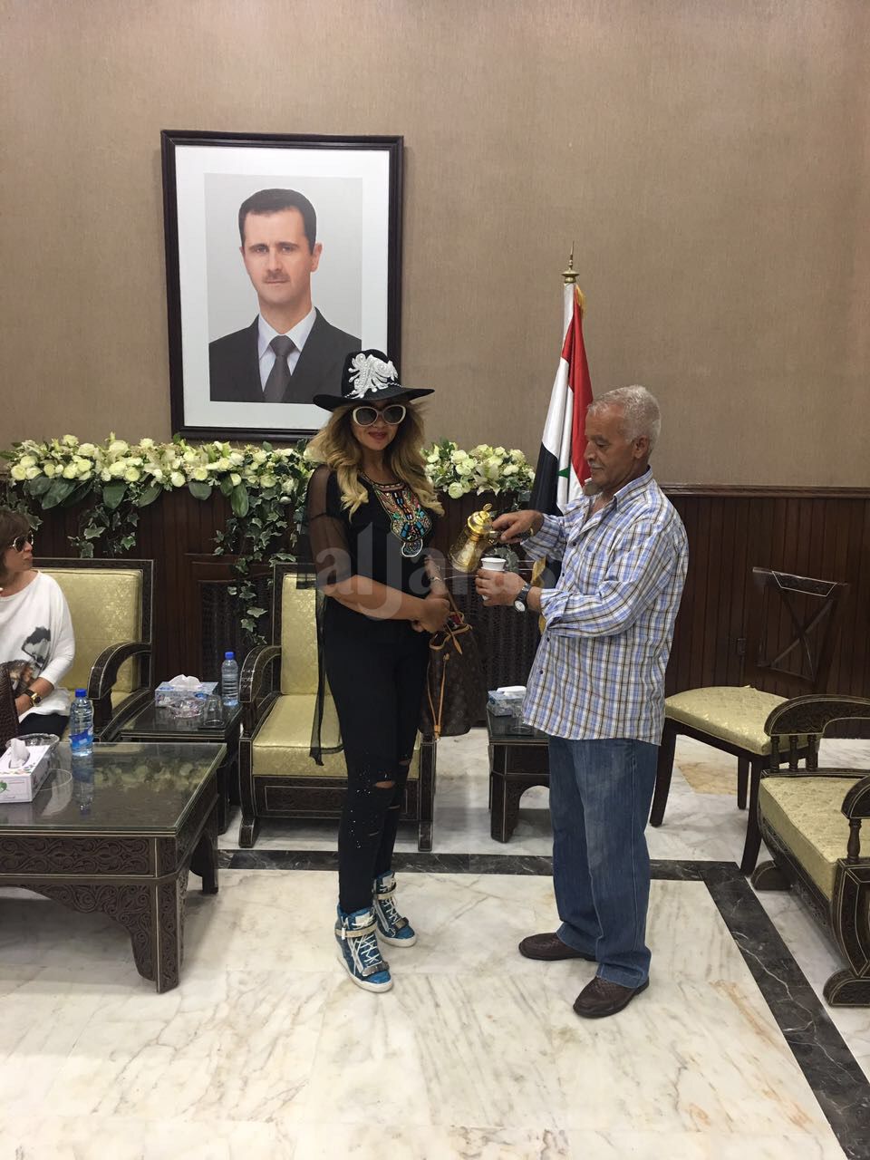 بوسي شلبي في سوريا وخلفه صورة الرئيس بشار الأسد